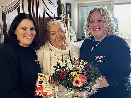 Die 1.500te Teilnehmerin des Hausnotrufs bekommt einen Blumenstrauß von zwei Mitarbeiterinnen überreicht