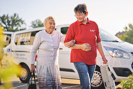 Eine Seniorin wird von einer ehrenamtlichen Begleitung vom Fahrzeug zur Tagespflegeeinrichtung begleitet und sicher am Arm gehalten.